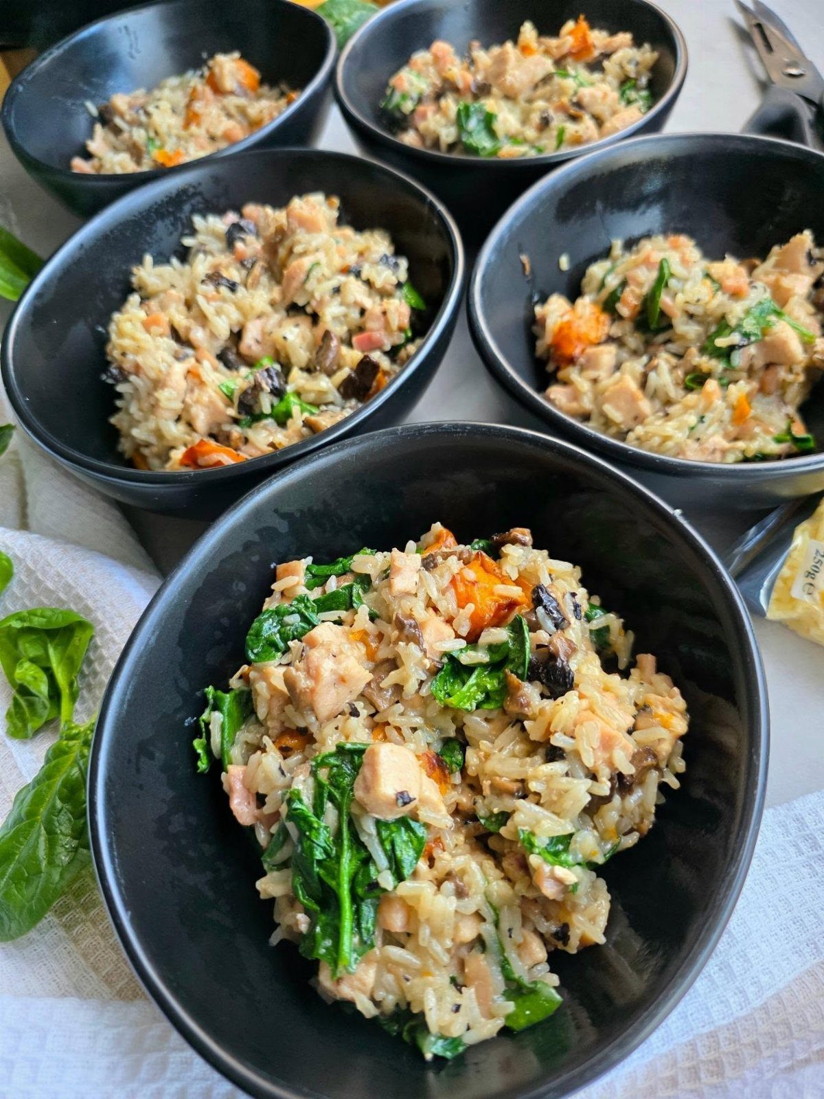 Creamy vegetable brown rice risotto served in five black bowls with fresh spinach.
