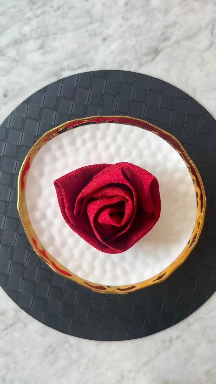 Red fabric napkin folded into a rose on a gold-rimmed white plate.