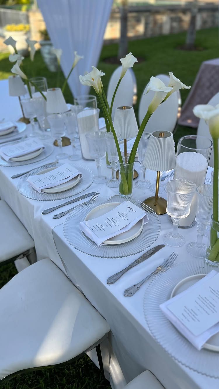 Fine dining table setting with white linens, calla lilies, and elegant glassware outdoors.
