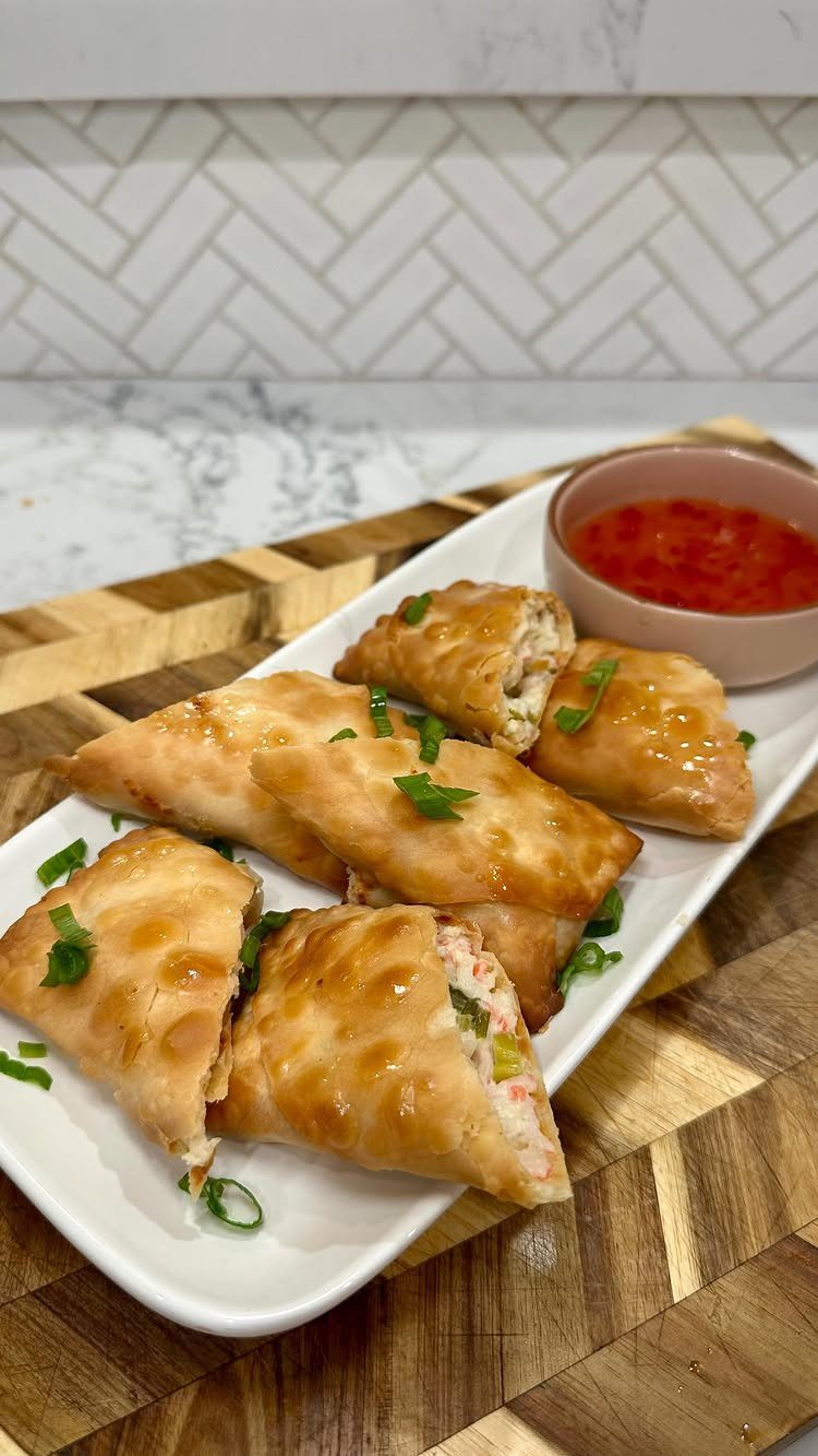 Crispy crab ragoons on a white platter with red dipping sauce and green onions.