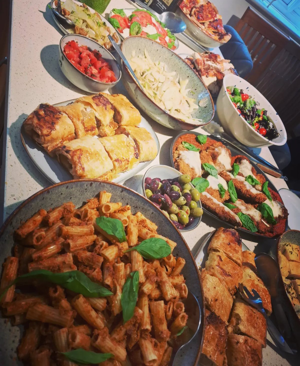Cheesy chicken breasts with basil alongside pasta and various side dishes on a table.
