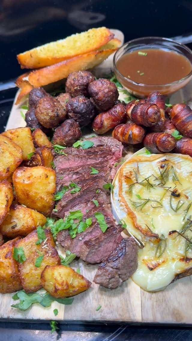 Sliced steak with roasted potatoes, carrots, melted cheese, and dipping sauce on wood.