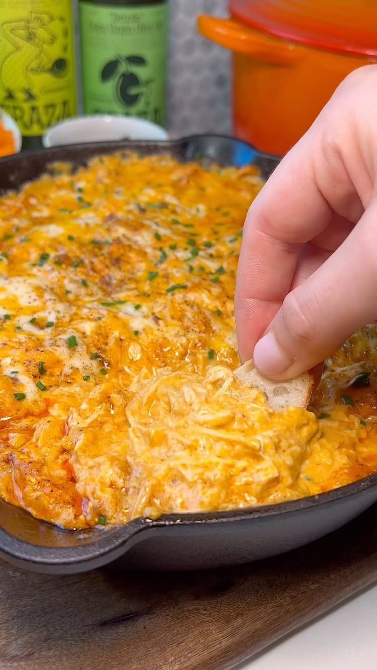 Creamy orange buffalo chicken dip in a cast-iron skillet with a bread dip.