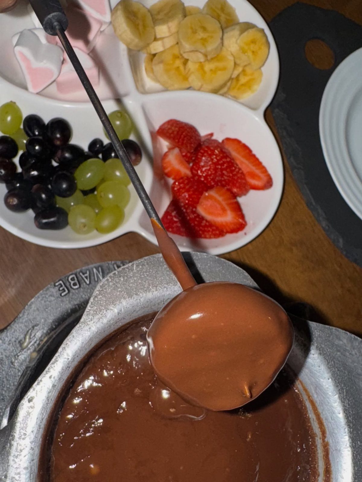 Chocolate fondue pot with a fork dipping food, surrounded by fresh fruit and marshmallows.