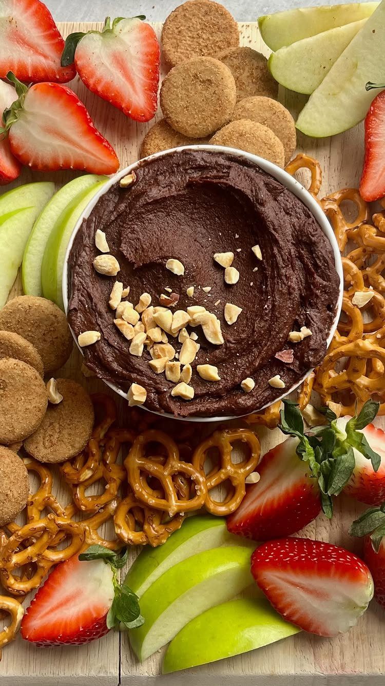 Chocolate hummus in a bowl surrounded by strawberries, apple slices, cookies, and pretzels.