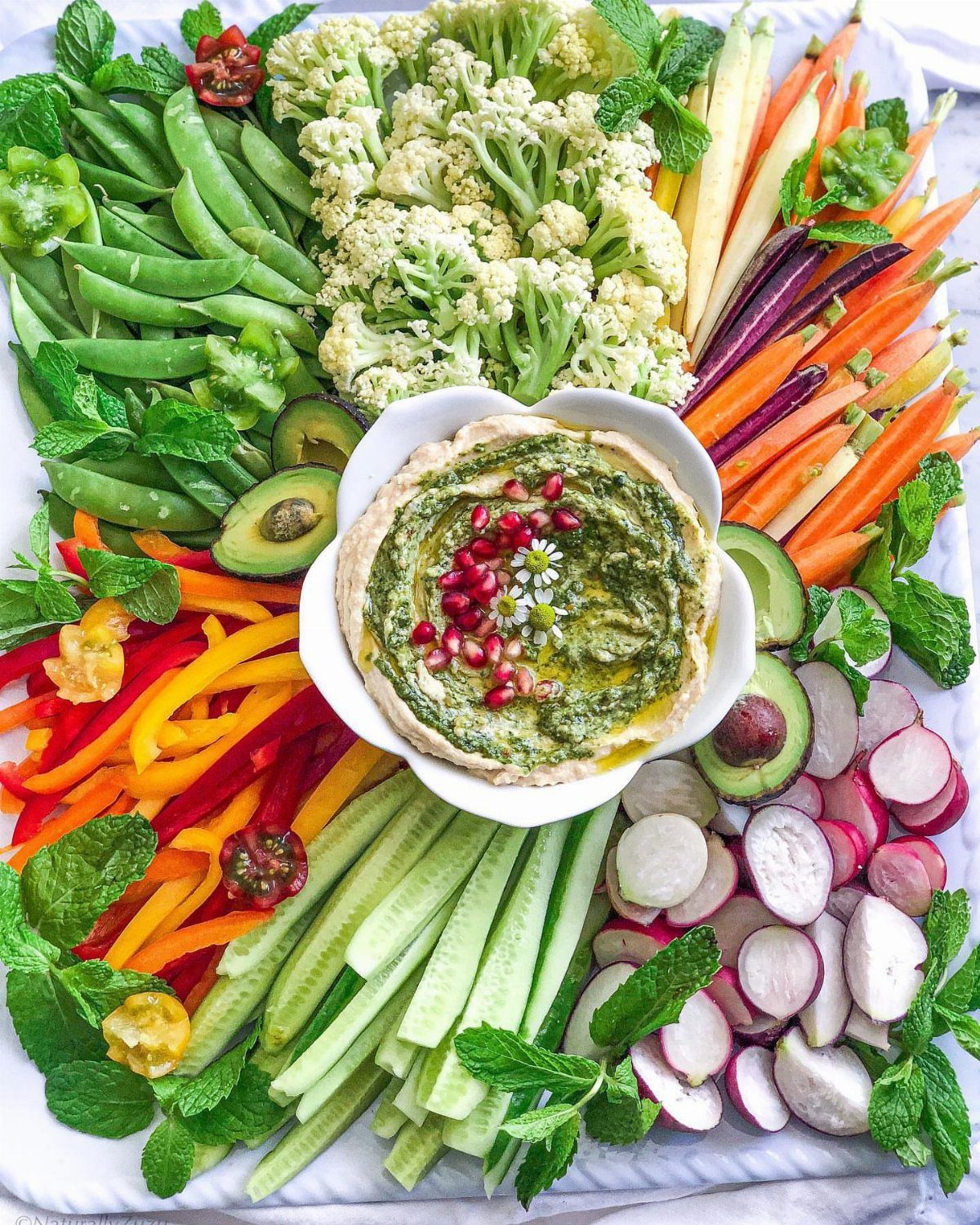 Green hummus with pomegranate seeds surrounded by a colorful variety of raw vegetables.