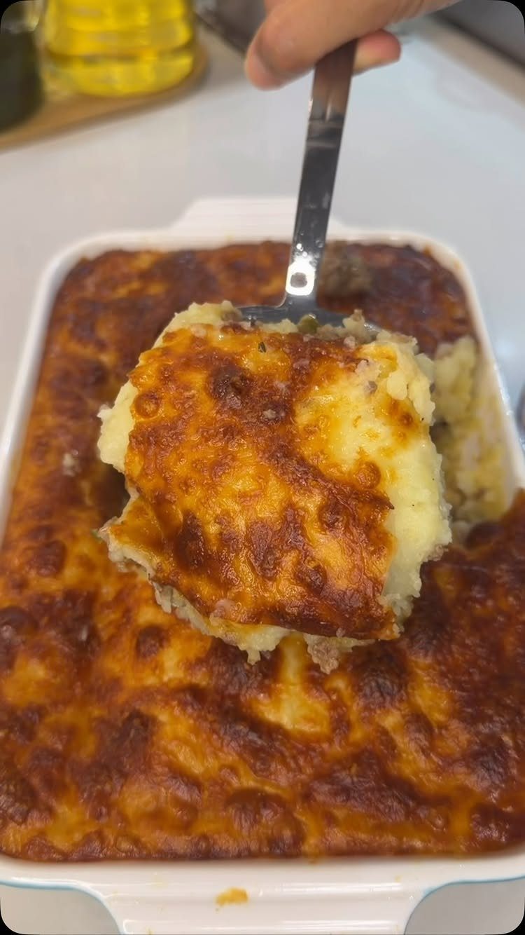 Cheesy mashed potato casserole being lifted from a white baking dish with a spatula.