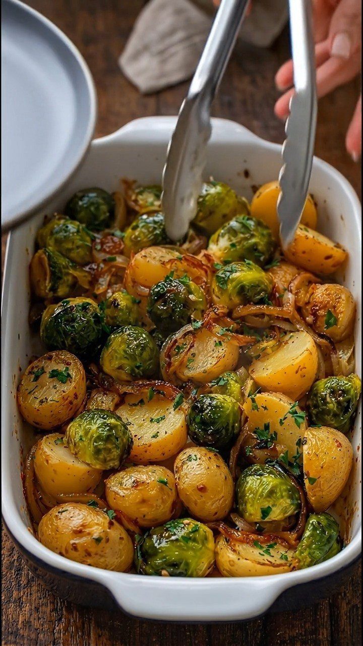 Roasted Brussels sprouts and golden potatoes in a white baking dish with tongs.