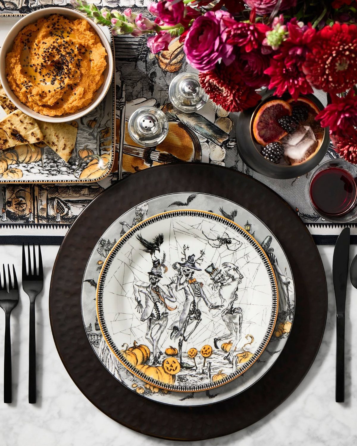 Skeleton illustrated dinner plate on a black charger with black cutlery and red flowers.