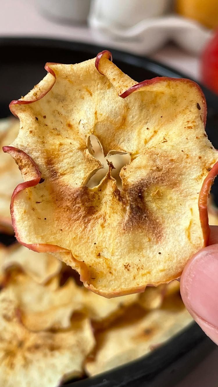 Dried apple chip with red skin held above a bowl of apple chips