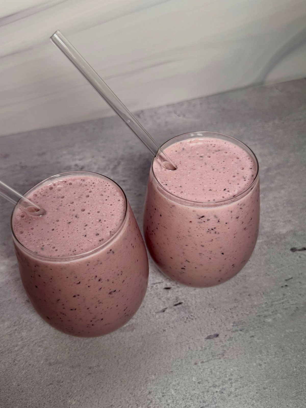 Two pink berry smoothies in glasses with straws on a grey surface.