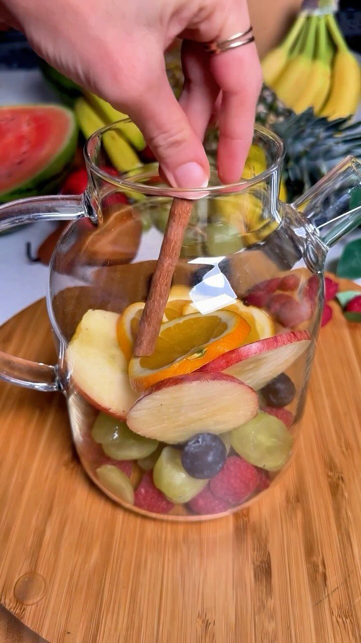 Glass pitcher of fruit-infused water with cinnamon stick, oranges, and berries on wood.