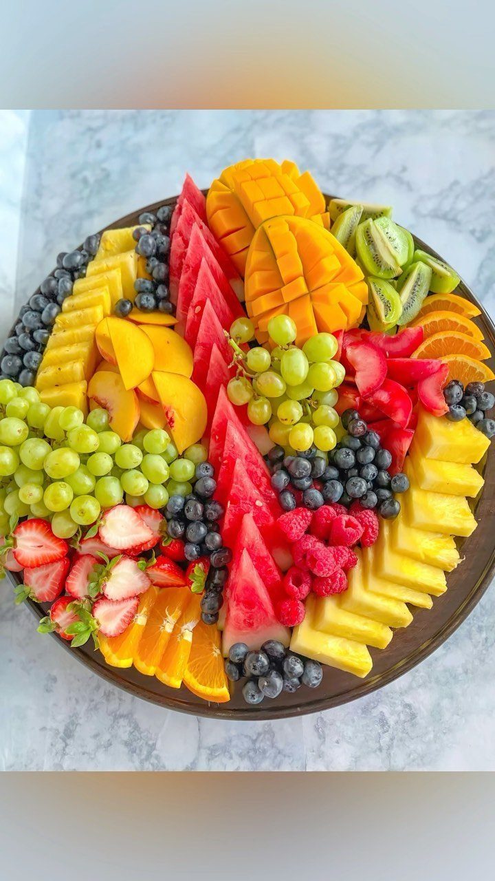 Fruit platter with sliced watermelon, pineapple, mango, kiwi, grapes, and berries on marble.