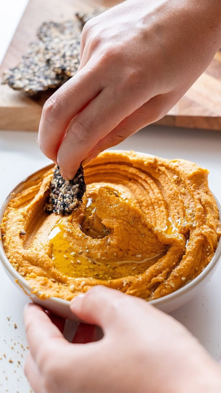 Orange hummus in a bowl with olive oil and a seedy cracker