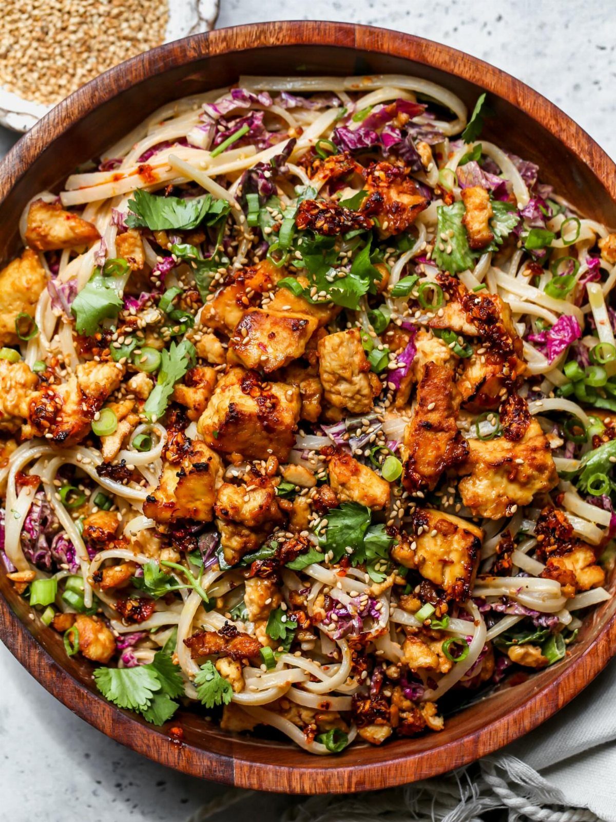 Noodles with tofu, purple cabbage, and cilantro in a wooden bowl topped with sesame seeds