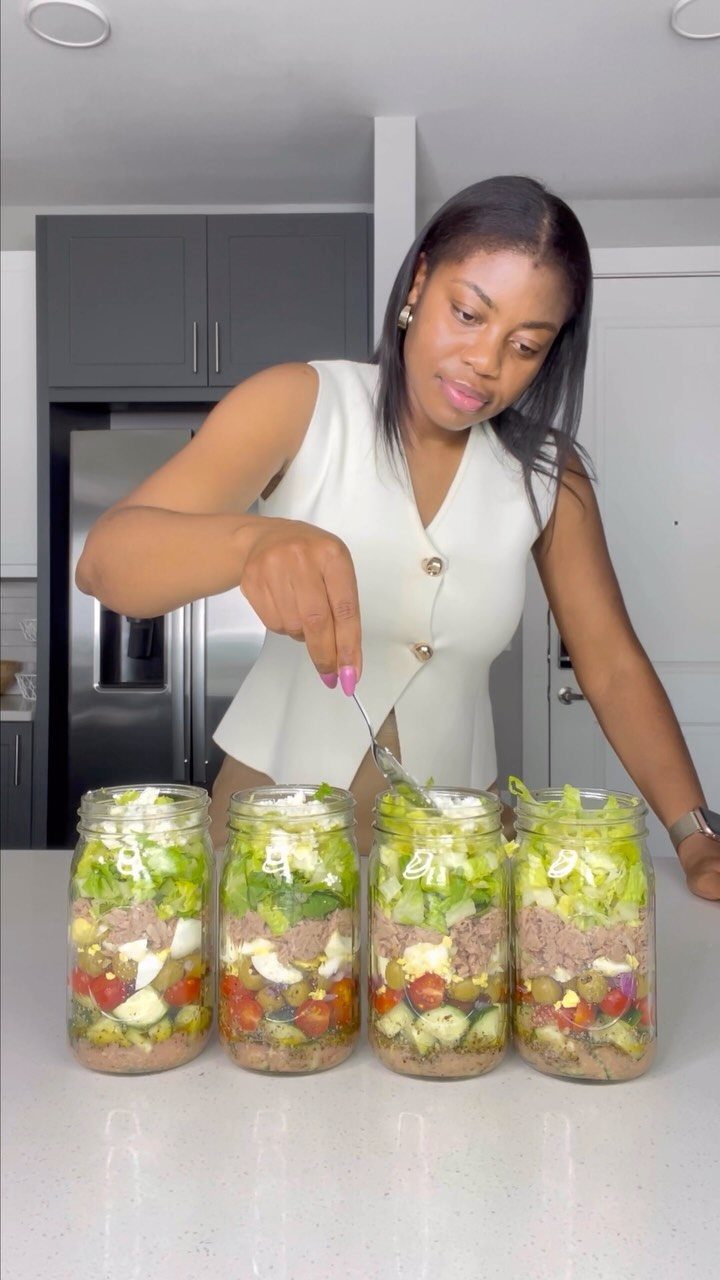 Woman preparing layered salads in four glass mason jars on a white kitchen counter.
