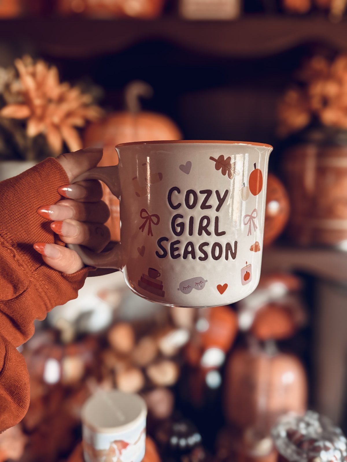 Beige ceramic mug with Cozy Girl Season text held against autumn background
