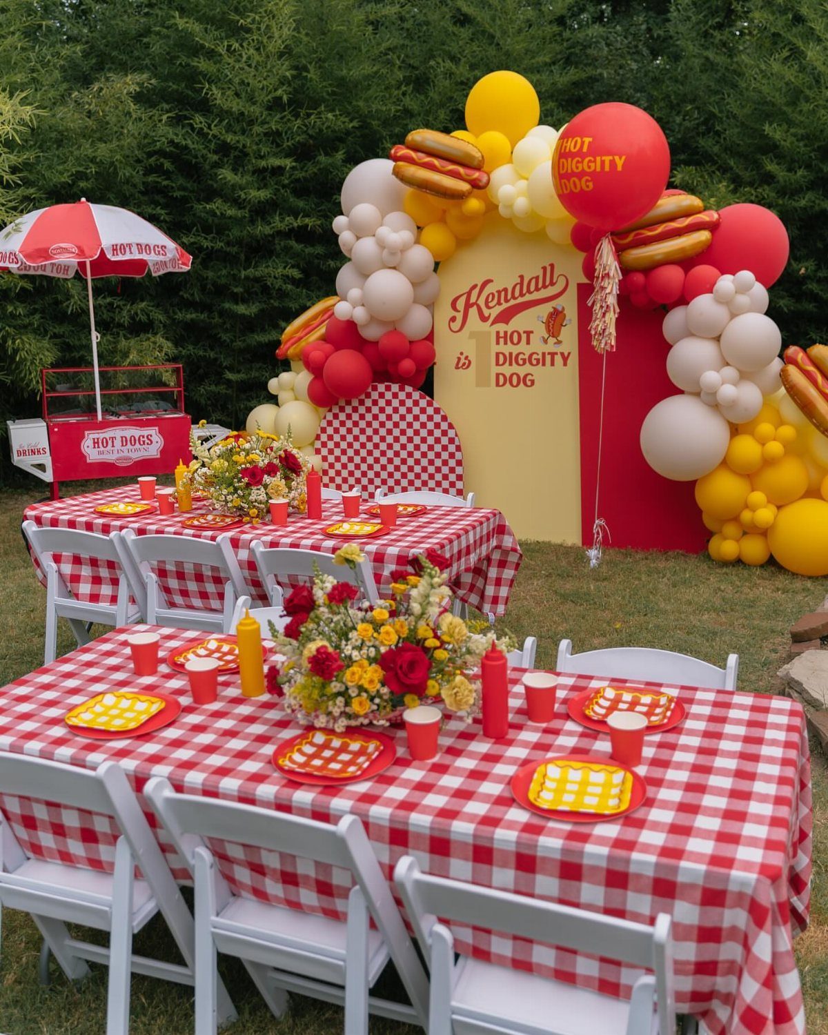 Outdoor hot dog party setup with checkered tables and a large balloon arch.