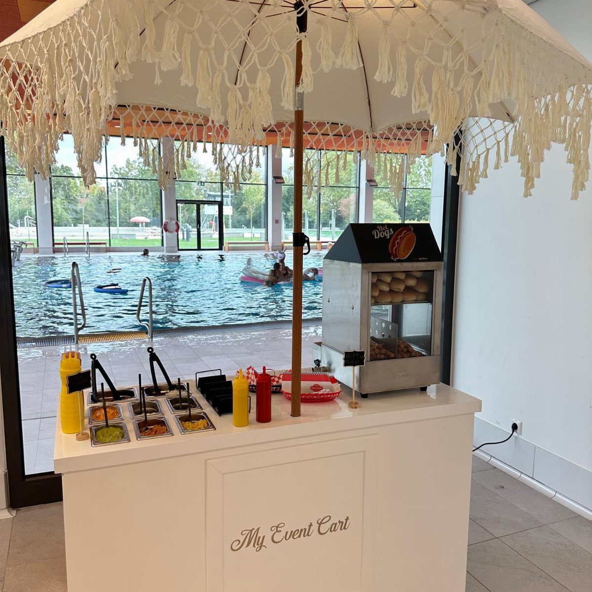 White event cart with hot dog steamer and condiments under a fringed umbrella.