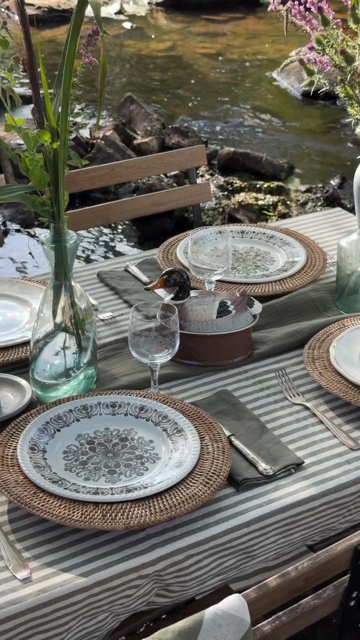 Outdoor table setting with woven chargers, patterned plates, and wine glasses by a river