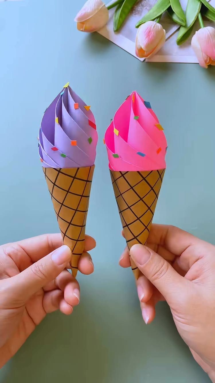 Paper ice cream cone crafts in purple and pink held by two hands.