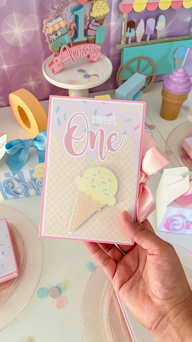 Hand holding a pink first birthday invitation featuring a yellow ice cream cone illustration.