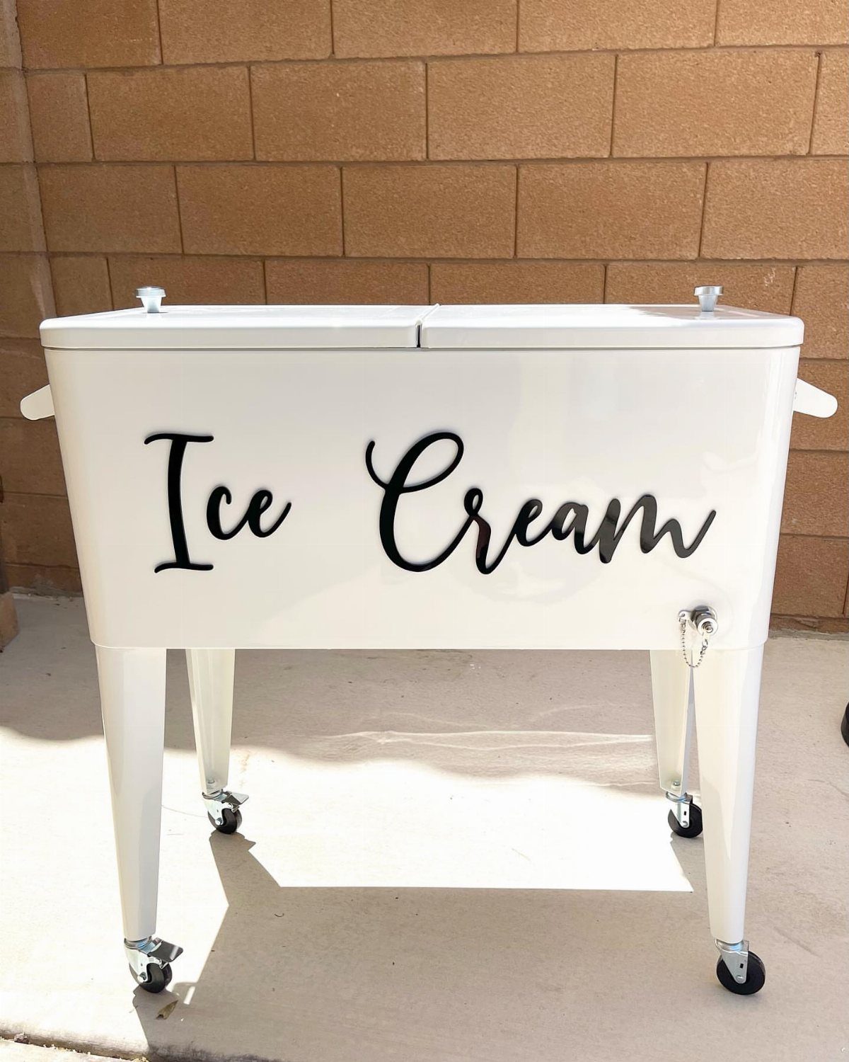 White vintage ice cream freezer cart with black cursive lettering against brick wall