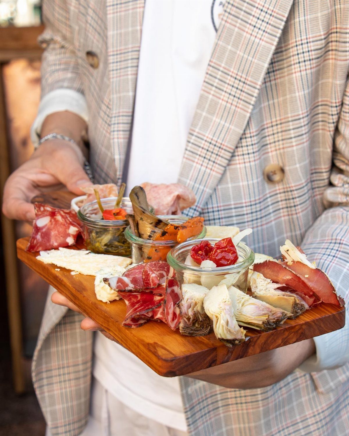 Antipasti platter with cured meats and vegetables on a wooden board held by a person.