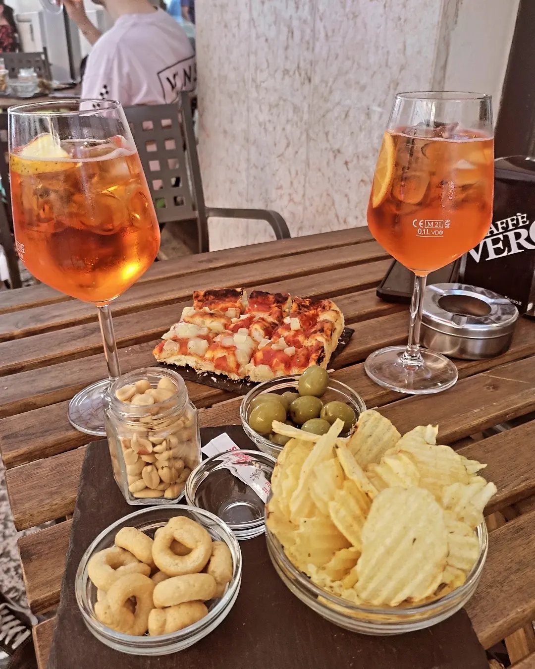 Two orange Aperol Spritz cocktails with bowls of chips, olives, and nuts.