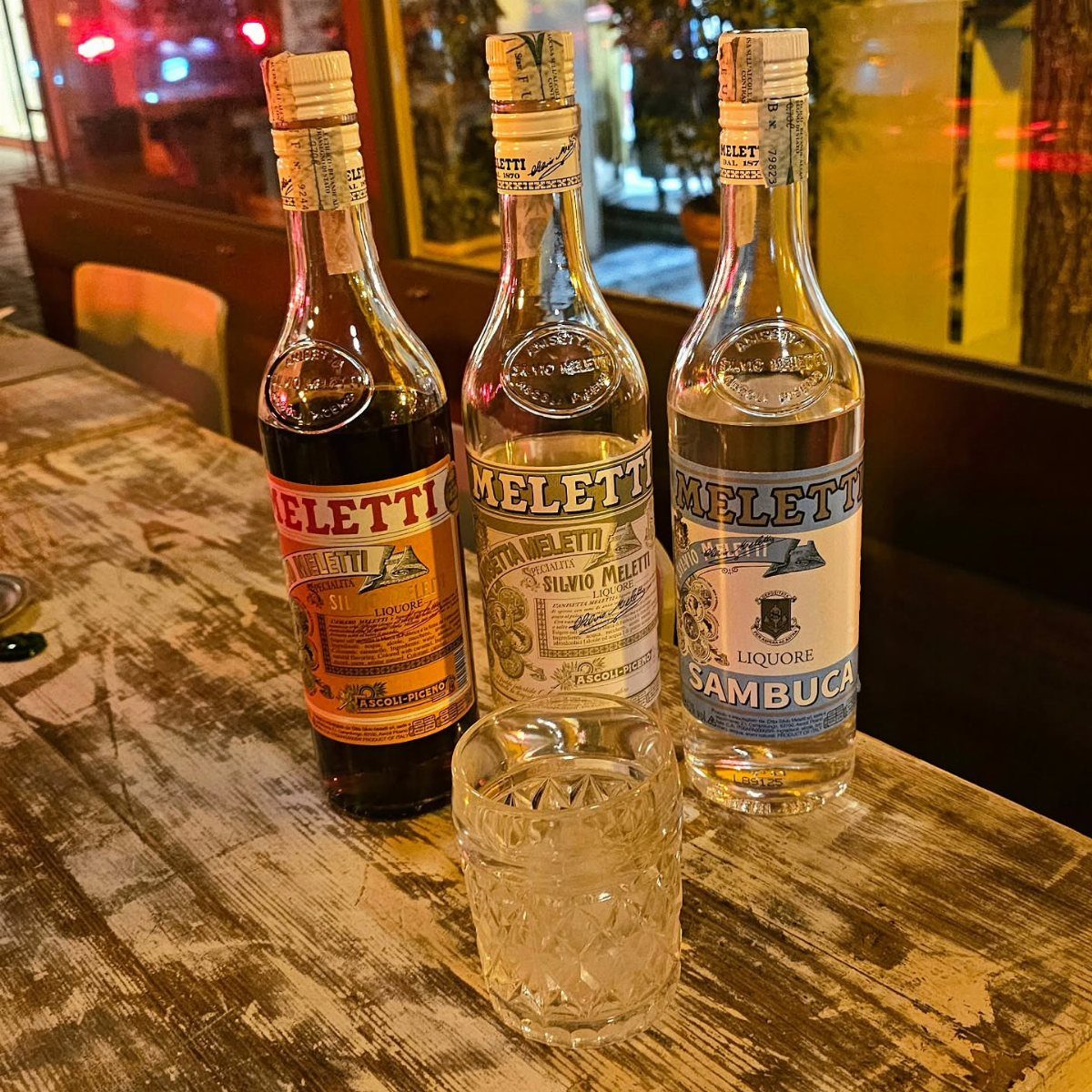 Three bottles of Meletti liqueur and a crystal glass on a wooden table.