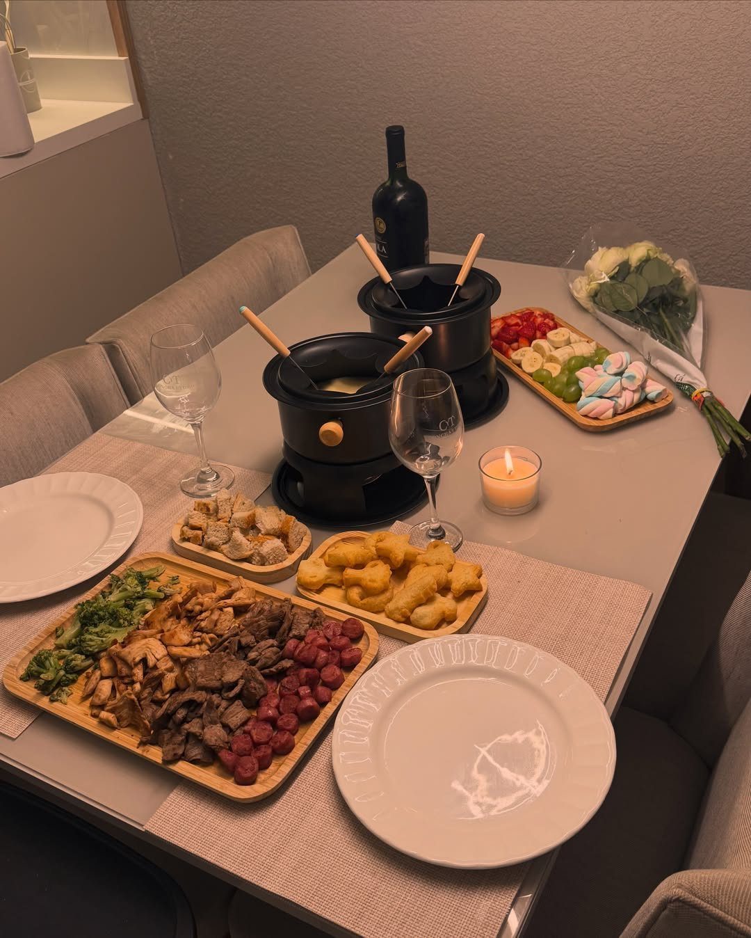 Two black fondue pots on a table with wine, meats, and vegetables.