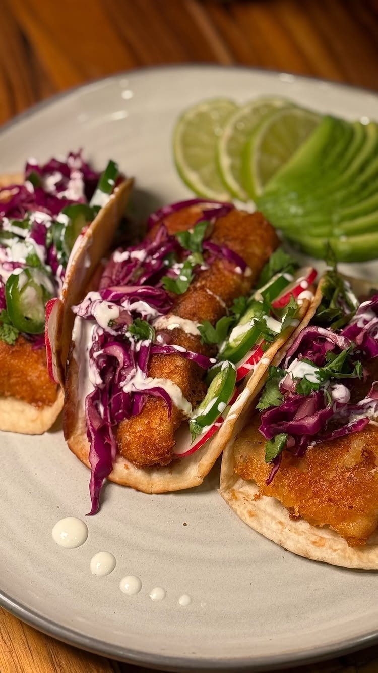 Fried fish tacos with purple cabbage and white sauce, served with lime and avocado.