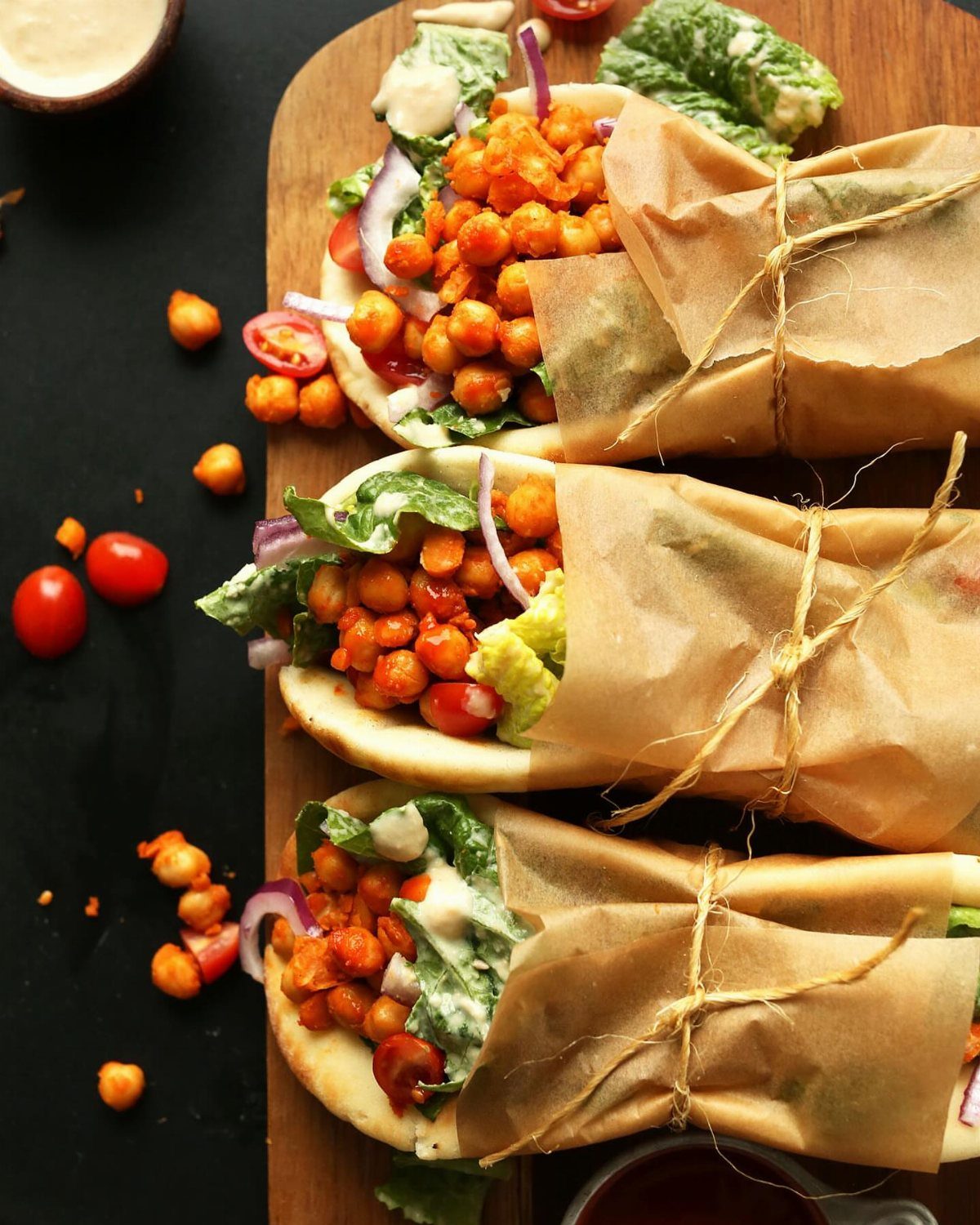 Three pita pockets filled with chickpeas and vegetables wrapped in brown parchment paper.