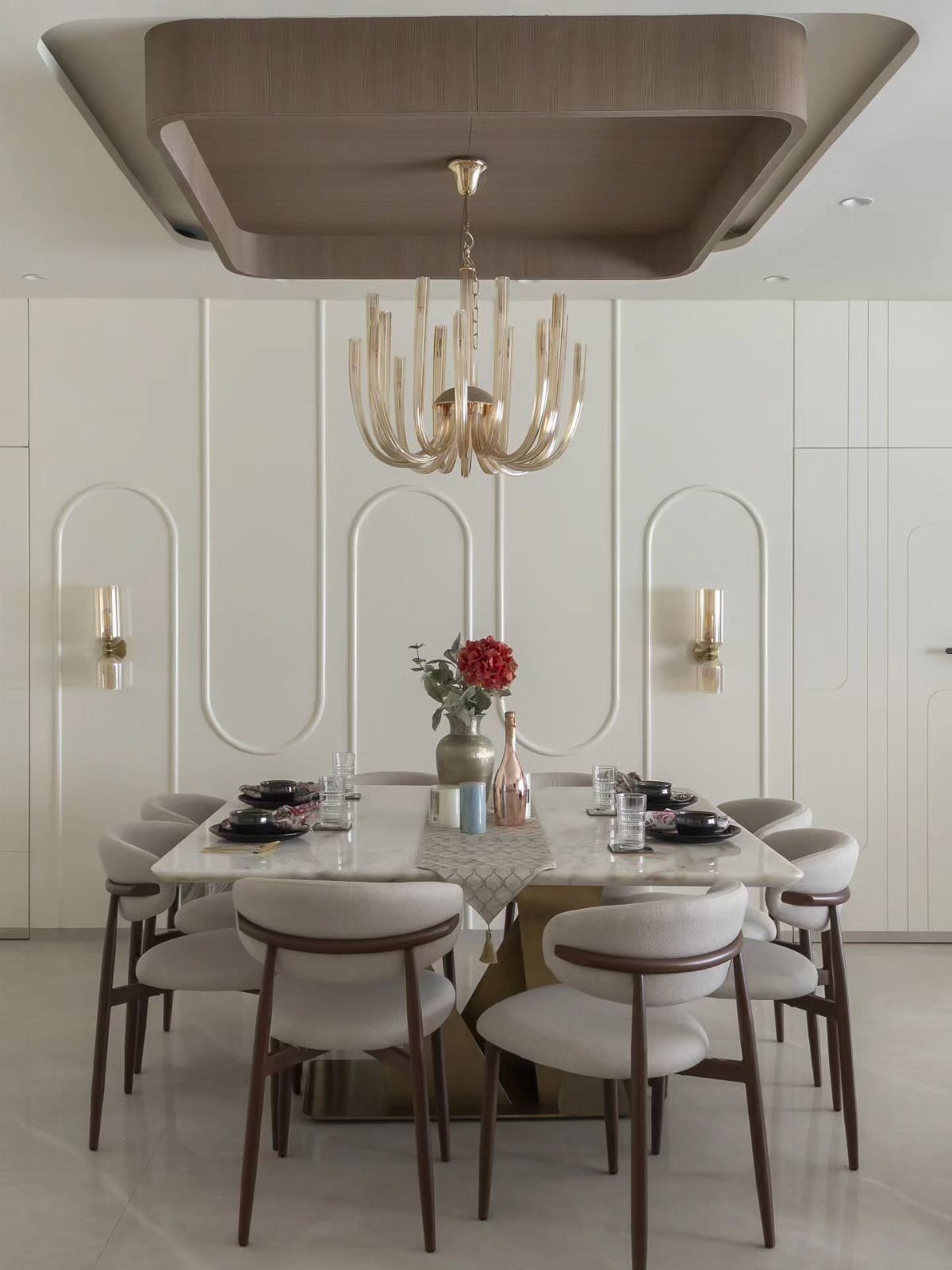Modern dining table with black plates, gold chandelier, and floral centerpiece in white room.