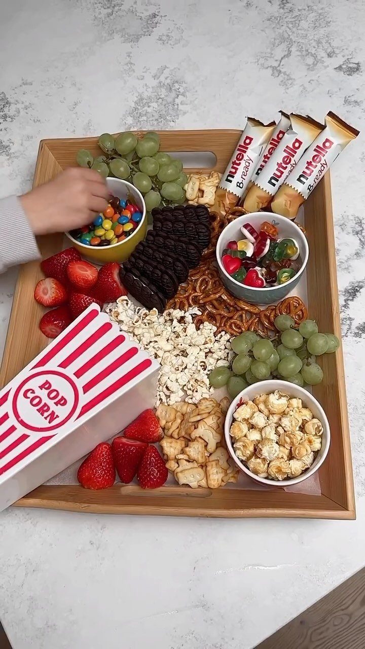 Wooden snack tray with popcorn, grapes, strawberries, chocolates, and candies for movie night.