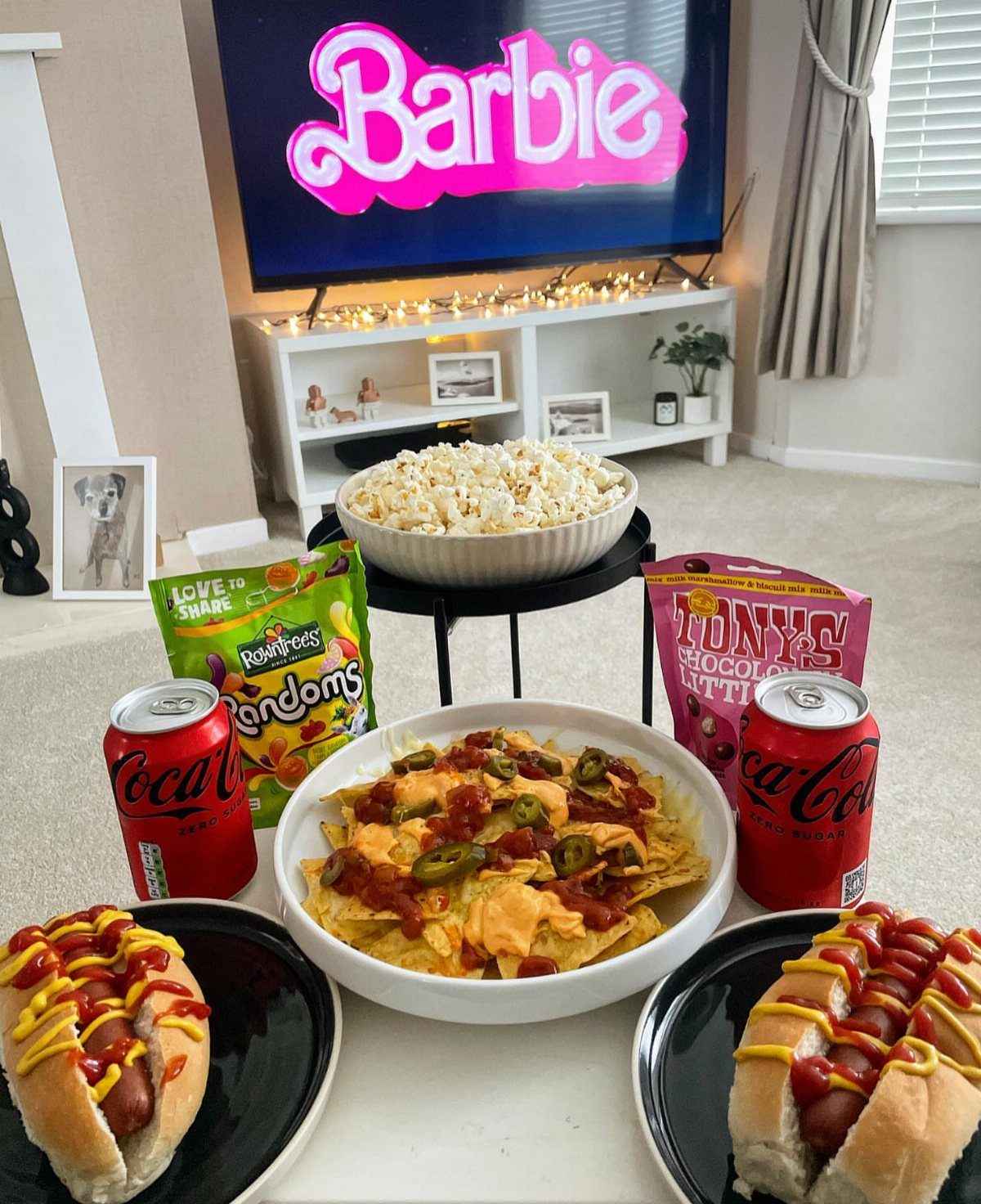 Movie snack spread with hot dogs, nachos, popcorn, and soda before a television.