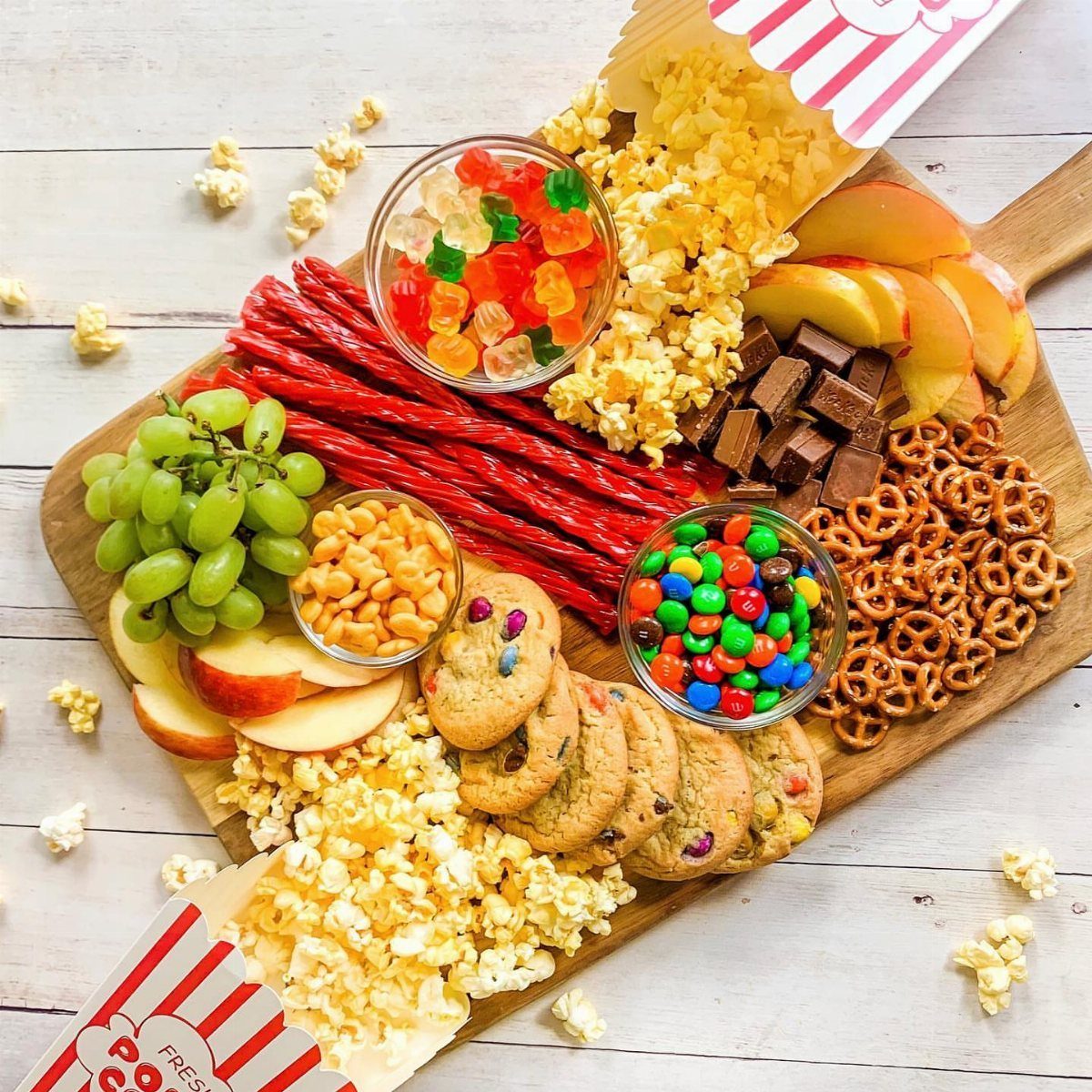 Assorted movie snacks including pretzels, popcorn, and fruit on a wooden serving board.