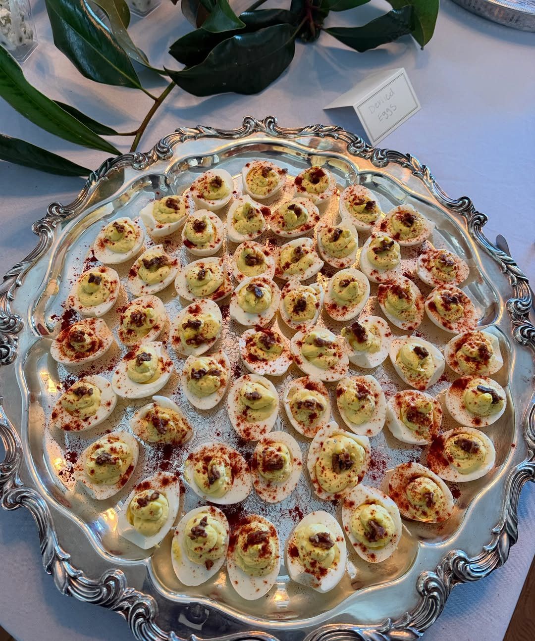 Deviled eggs with paprika on an ornate silver platter over a white tablecloth.