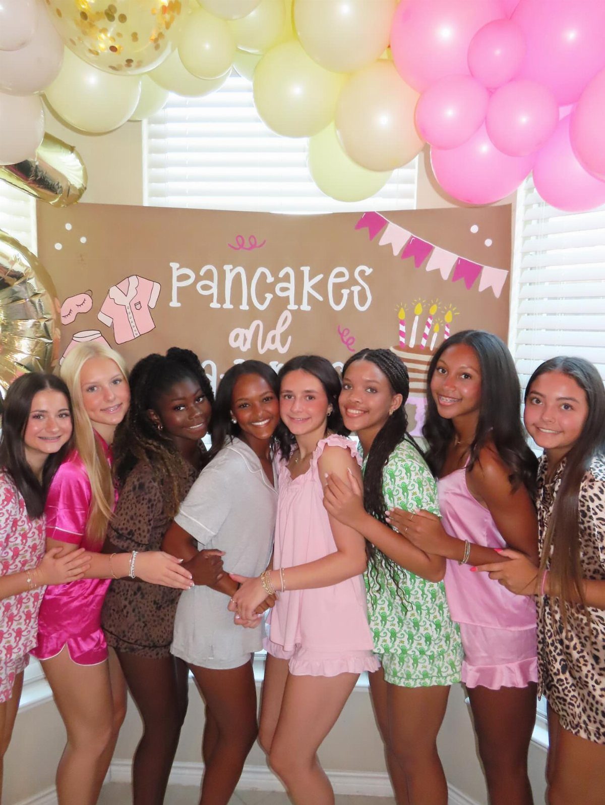 Eight teenage girls in colorful pajamas posing in front of a party banner.