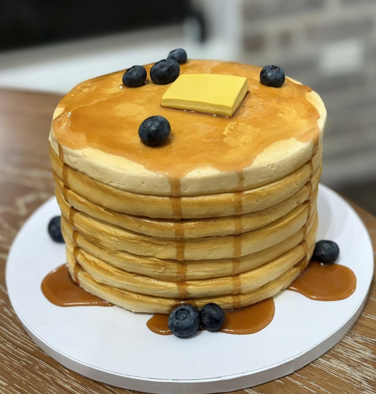Pancake cake topped with butter and blueberries, drizzled with syrup on white board.