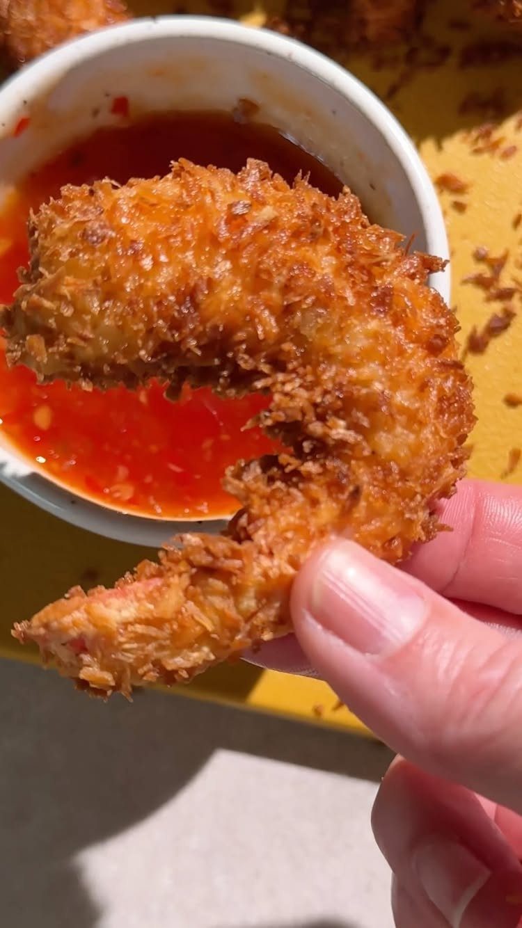 Coconut breaded fried shrimp held over a bowl of red dipping sauce.