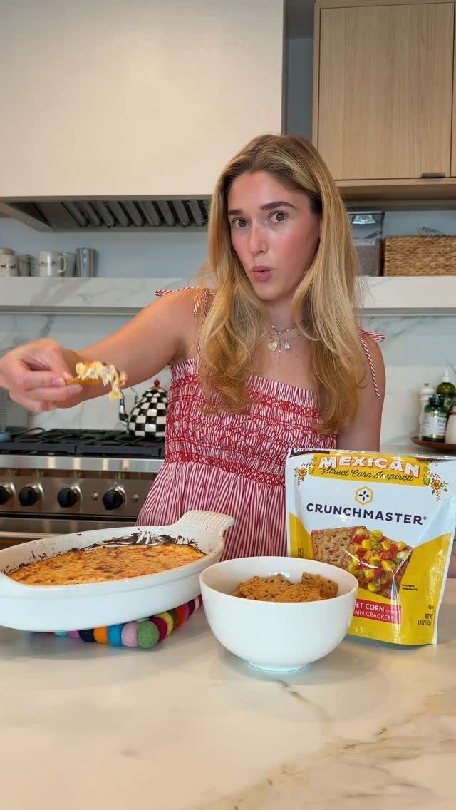 Woman holding a chip with corn dip next to a baking dish and chips.
