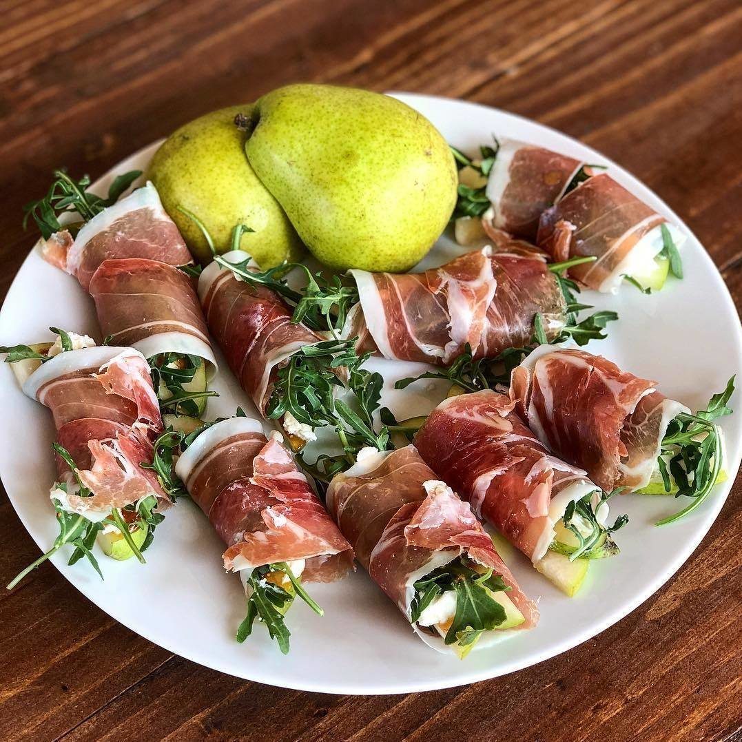 Prosciutto-wrapped pear slices with arugula and cheese on a white ceramic plate.