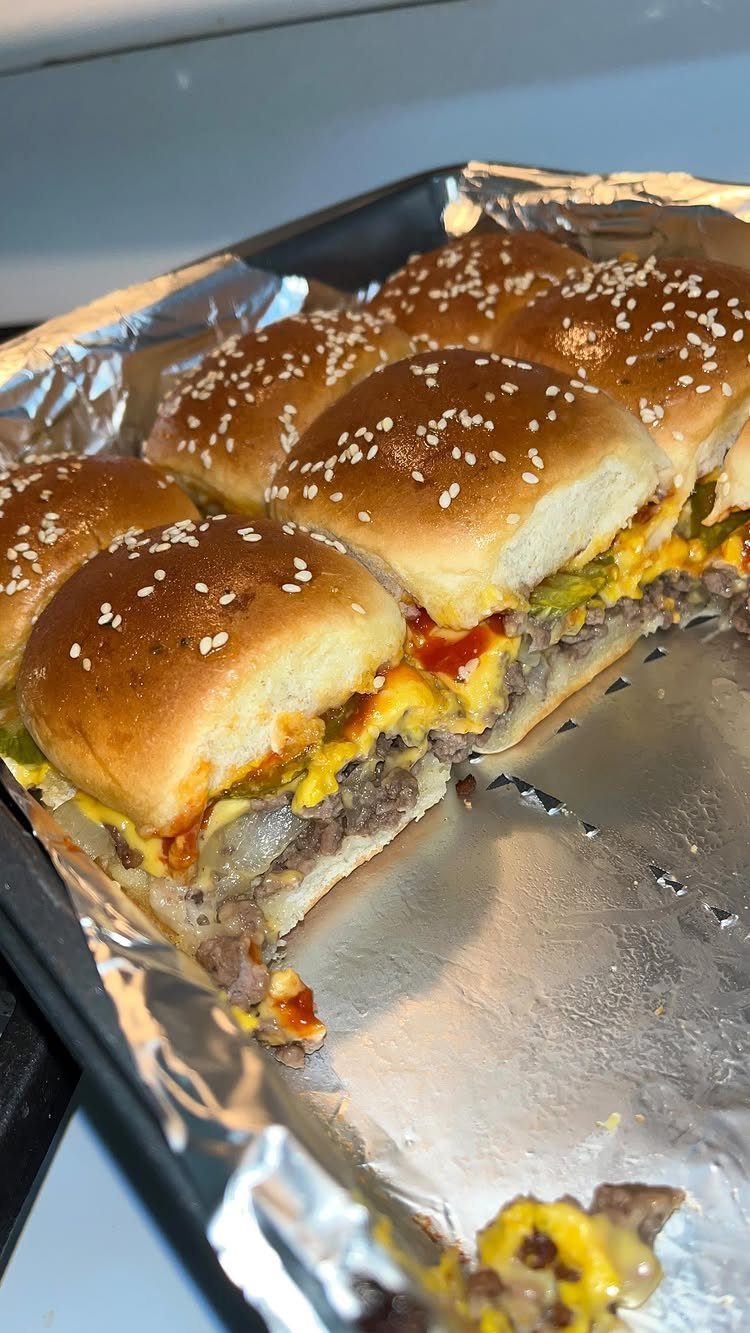 Mini cheeseburger sliders with melted cheese on sesame buns in a foil-lined tray.