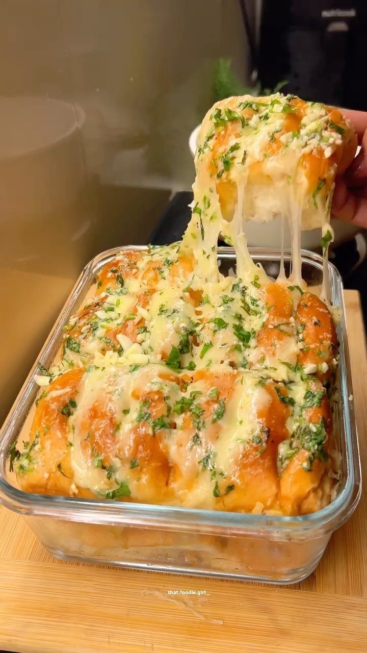 Cheesy dinner rolls with herbs in a glass dish, one being pulled away.
