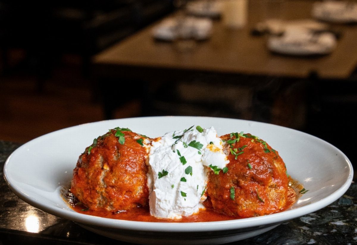 Two meatballs in red sauce with ricotta cheese and parsley in a white bowl.