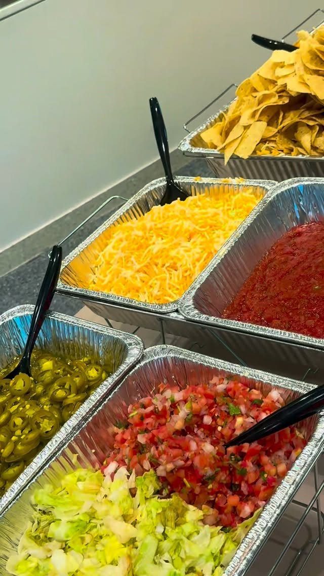 Taco bar toppings in foil trays including lettuce, salsa, cheese, and tortilla chips.