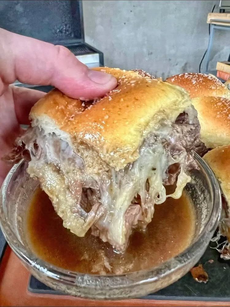 Cheesy beef slider being dipped into a bowl of brown au jus gravy.