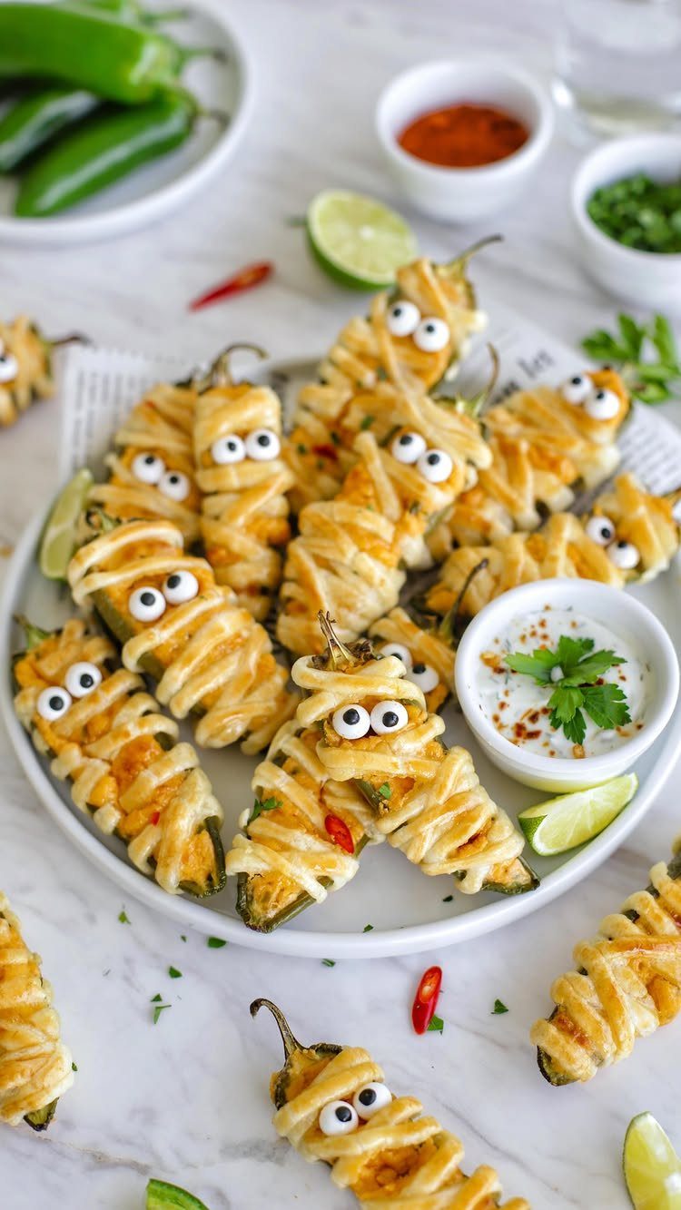 Mummy-themed stuffed jalapeno peppers with cheese drizzle and candy eyes on a plate.