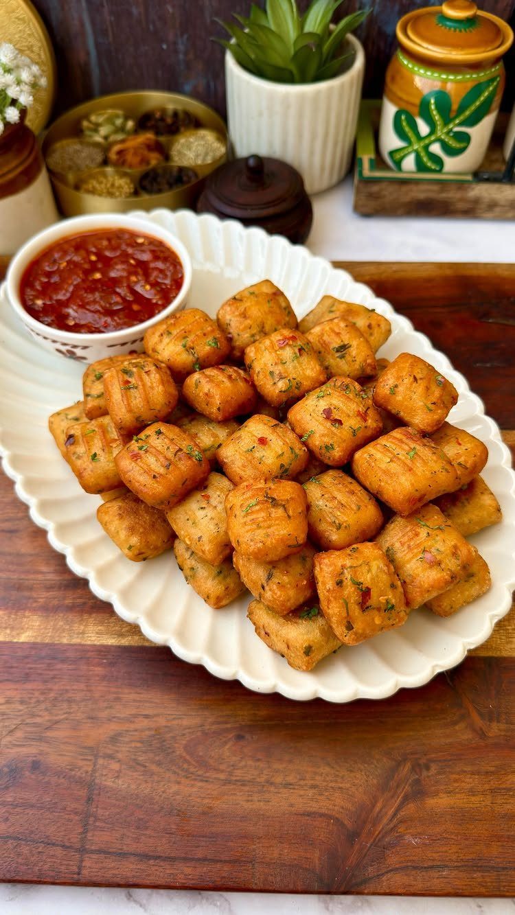 Golden-brown cubed spiced potato bites on a white platter with red dipping sauce.